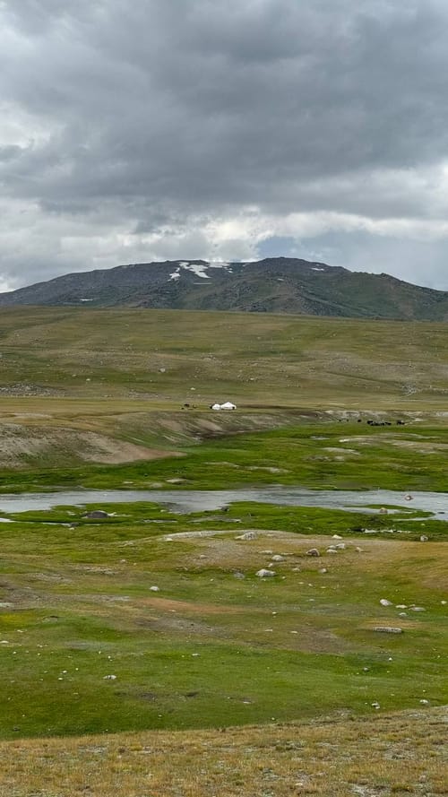 A cloudy hillside with distant gers and a grazing herd beside a river